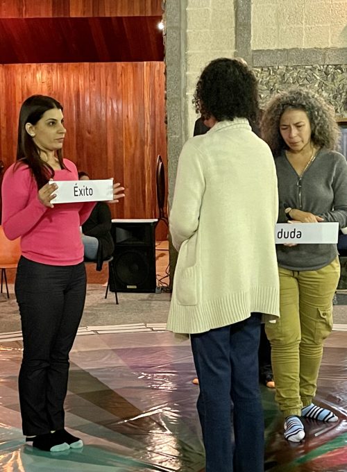 Mujer sostiene un letrero con un sentimiento, mientras otras 2 lo miran. Todo sobre una rueda de colores que representan el mundo emocional de una persona que esta parada en el centro.