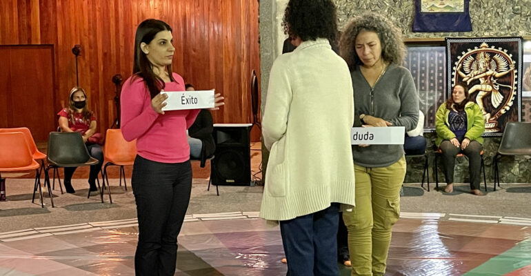 Mujer sostiene un letrero con un sentimiento, mientras otras 2 lo miran. Todo sobre una rueda de colores que representan el mundo emocional de una persona que esta parada en el centro.